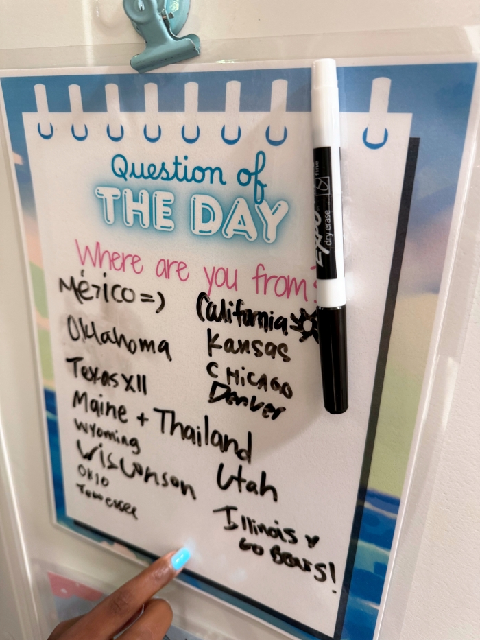 Close-up of a laminated “Question of the Day” sign on a Disney Cruise stateroom door. The question, written in pink, reads “Where are you from?” with handwritten responses like Mexico, Oklahoma, Thailand, and Illinois. A dry erase marker is clipped to the sign, and a finger with blue nail polish points to an answer.